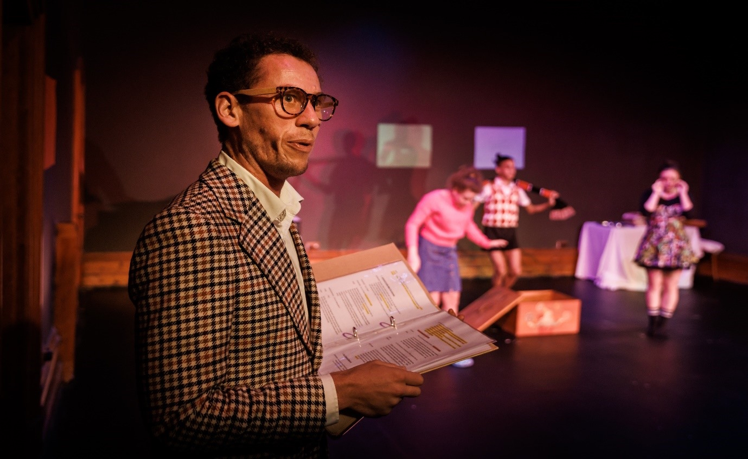 VV's audio describer Josh Lynzaat stands on stage wearing a tweed jacket, glasses, holding open book with written descriptions. In background on stage also but out of focus, three young people perform. With permission from La Mama for Kids Meet the Grandies production directed by Amelia Burke. Photo by Darren Gill